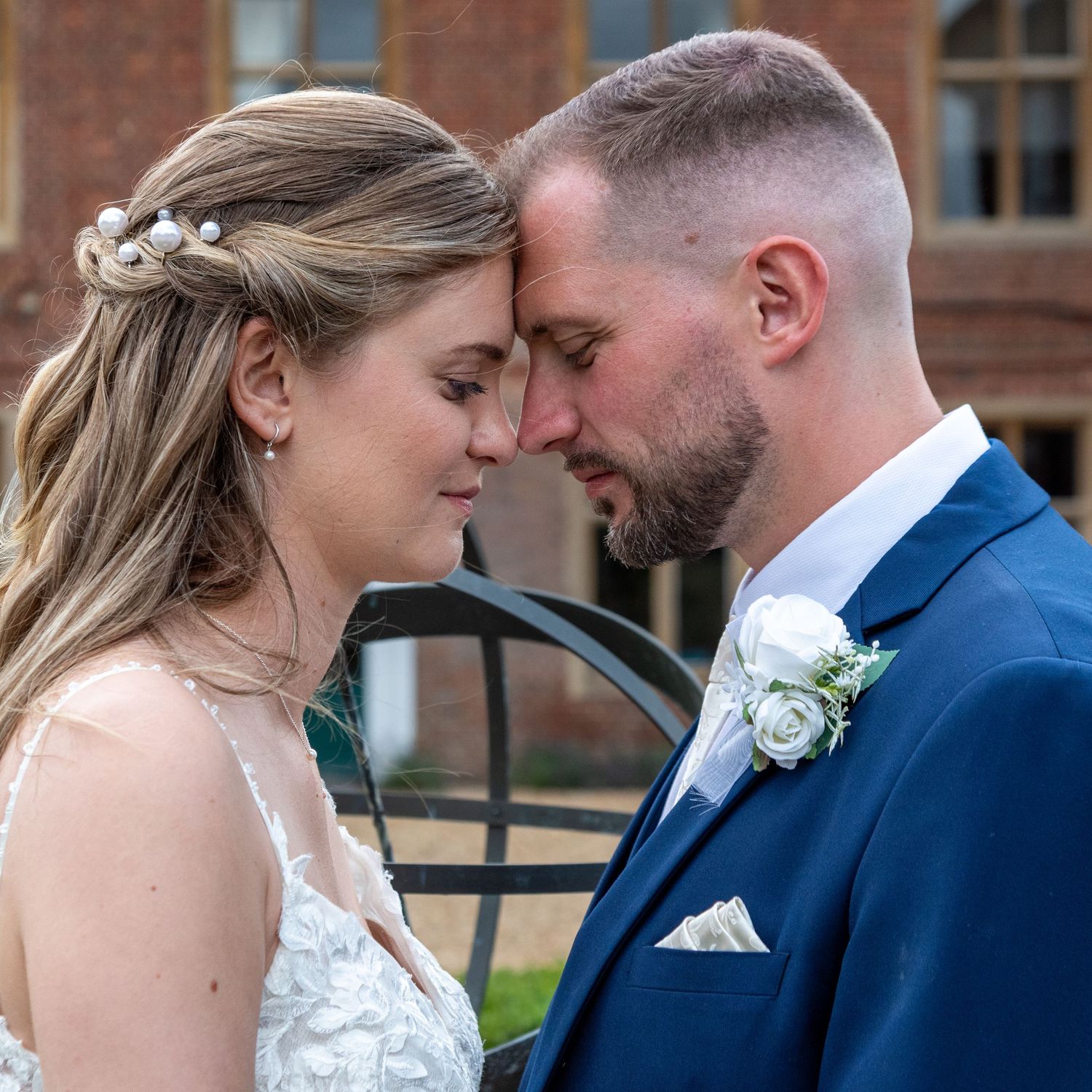 A bride and groom share an intimate forehead touch moment, dressed in wedding attire with a brick building backdrop.