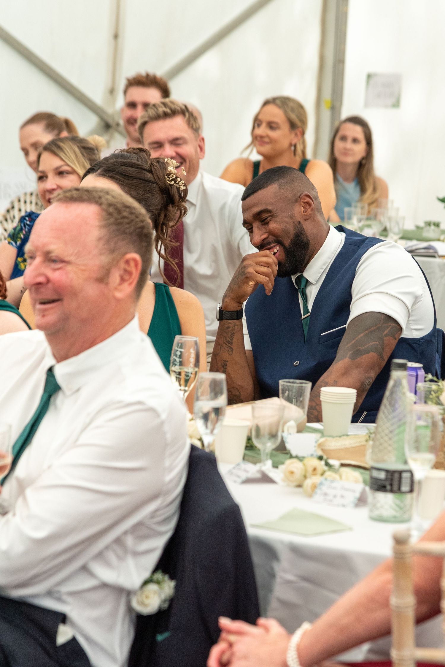 Guests laughing and enjoying speeches at an outdoor wedding reception under a marquee tent.