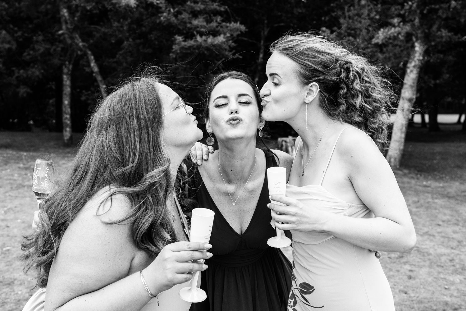 Three women celebrating with champagne glasses, two kissing the middle woman on cheeks, black and white outdoor photo.