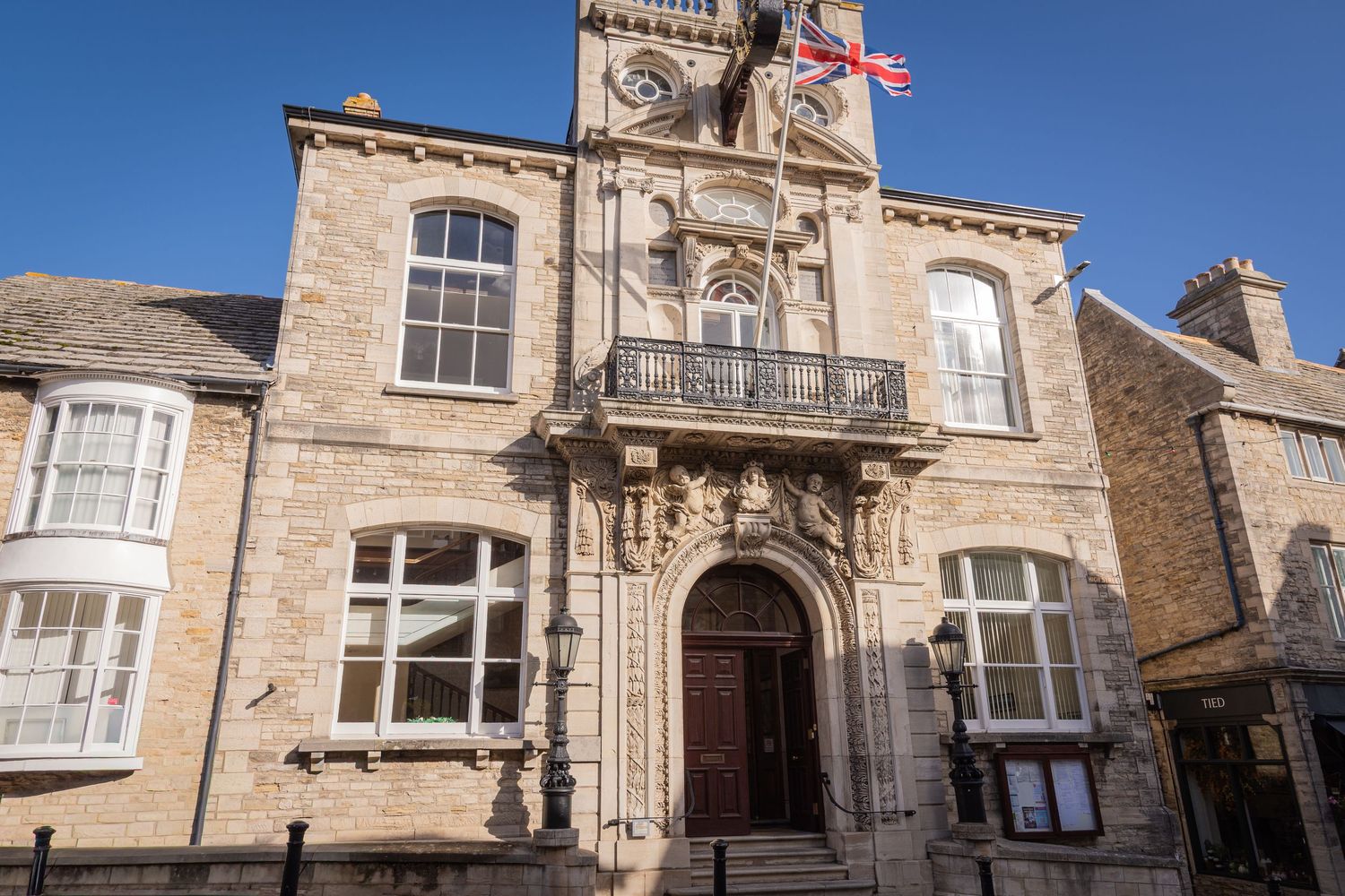 Stone buildings with ornate architectural details and a Union Jack flag flying against a bright blue sky in England.