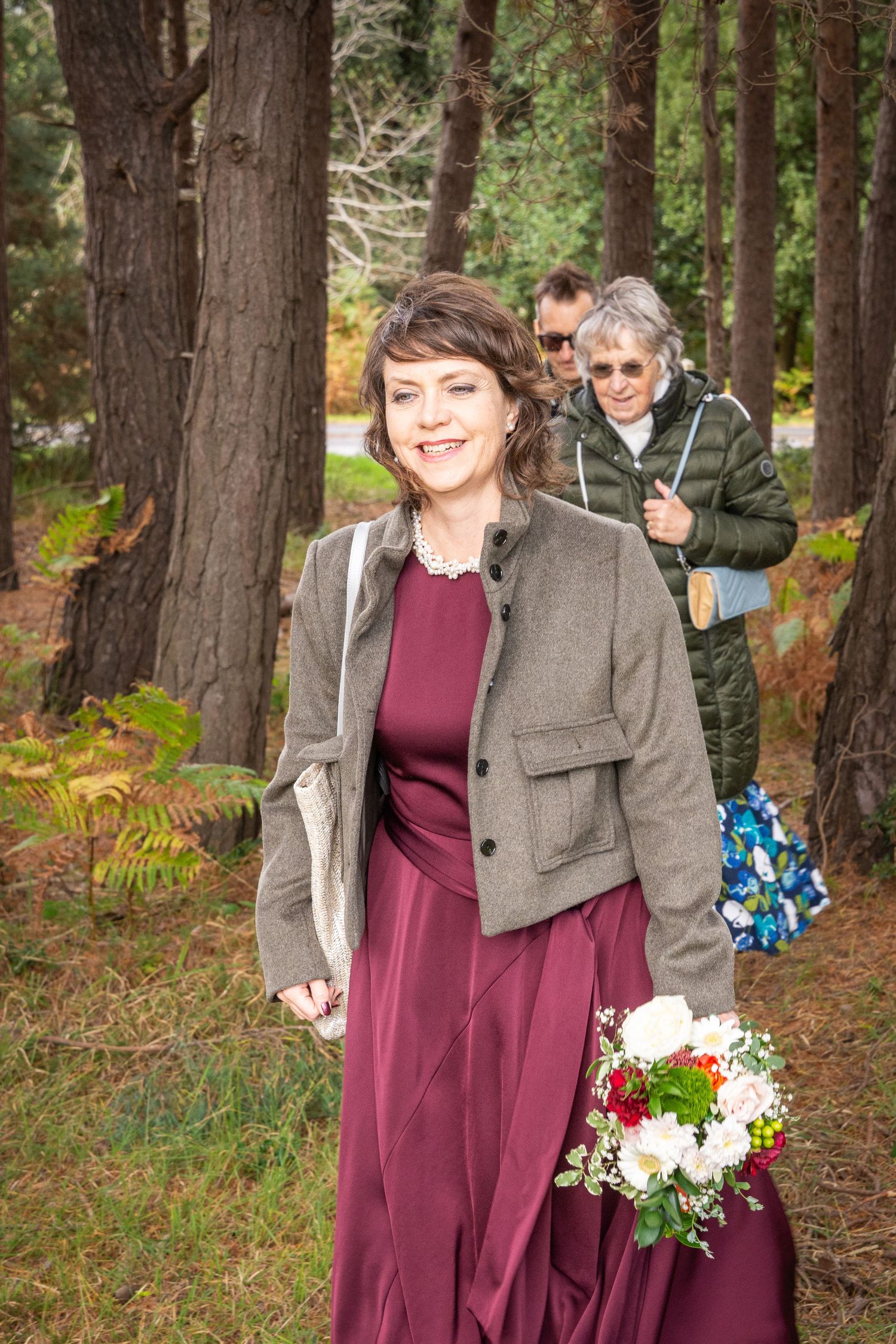 A couple poses together in a wooded forest setting wearing burgundy and gray attire with flowers.