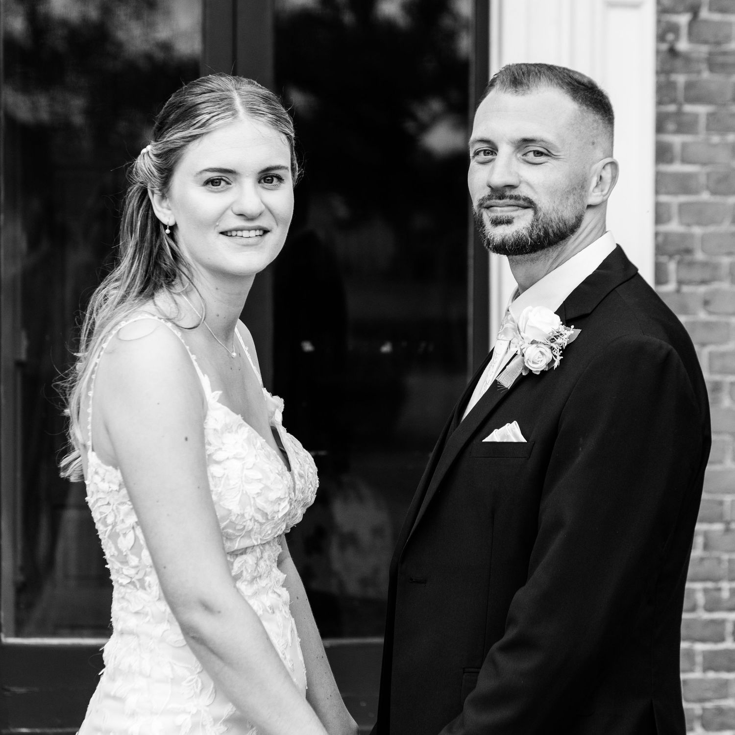 Black and white portrait of an elegant bride and groom posing together outside a brick building on their wedding day.