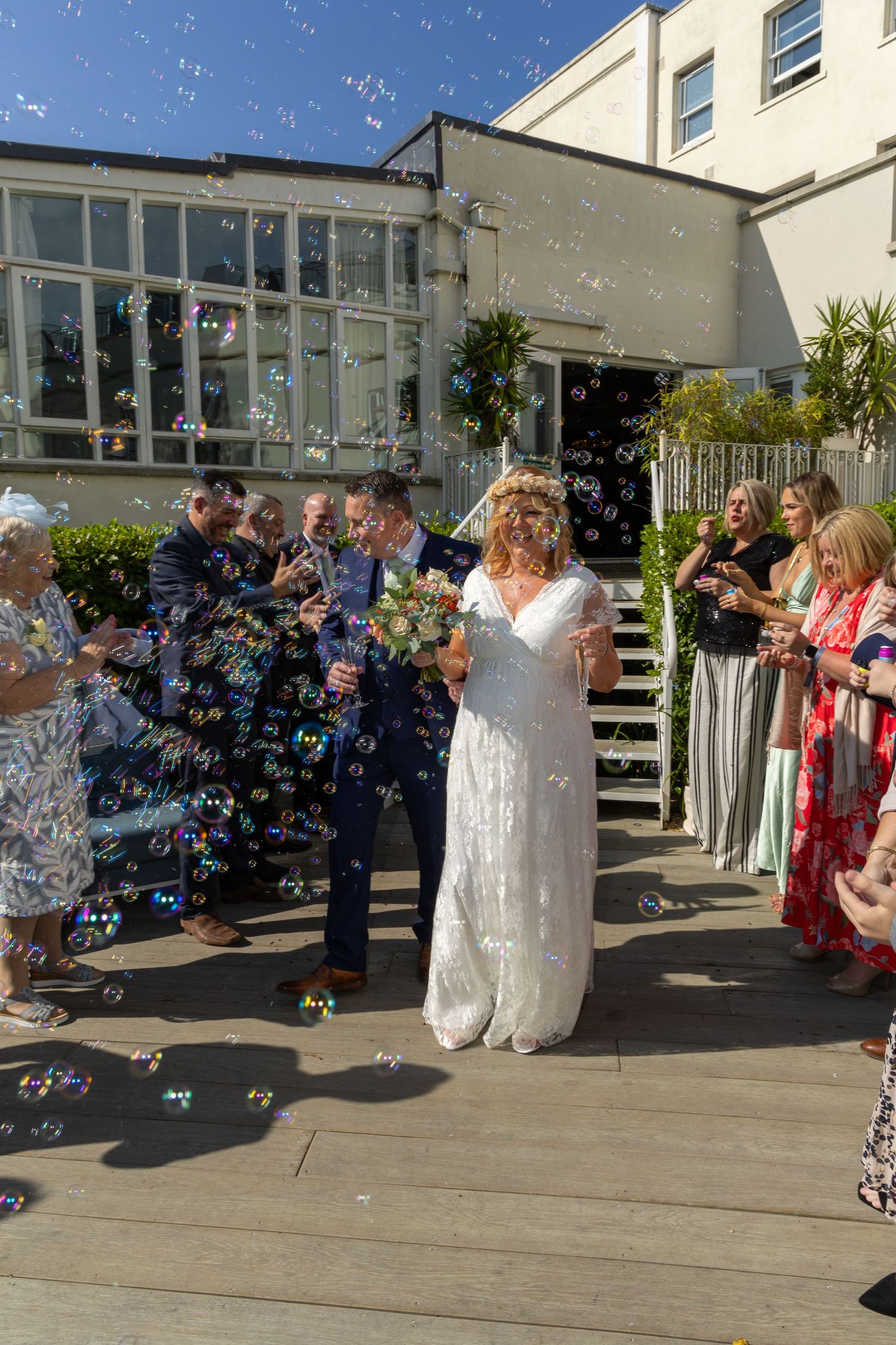 Bride and groom walk through bubble shower surrounded by smiling wedding guests outdoors on sunny day.