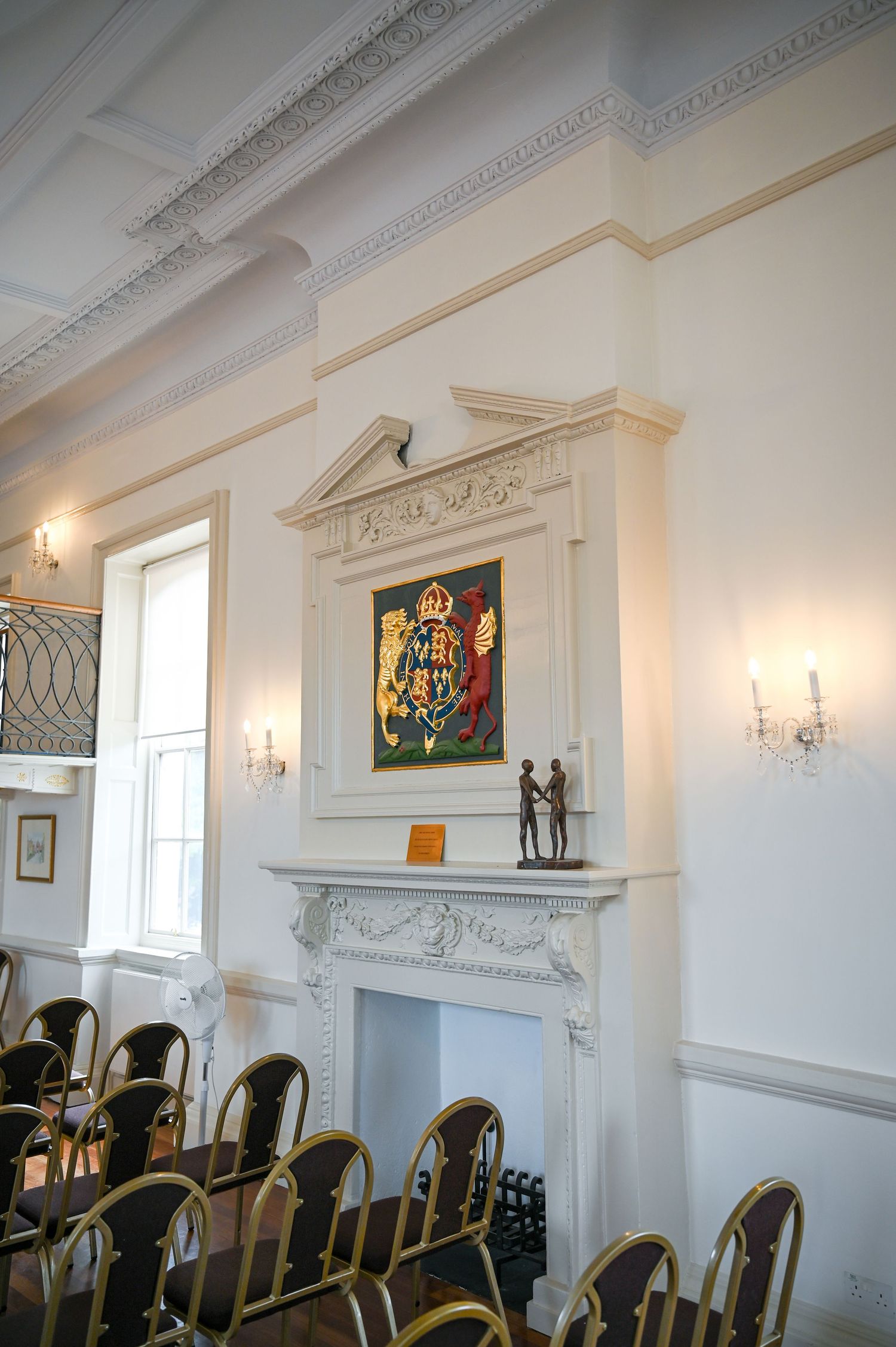 Elegant Georgian ceremony room with ornate fireplace, royal coat of arms artwork, wall sconces, and rows of gold chairs.