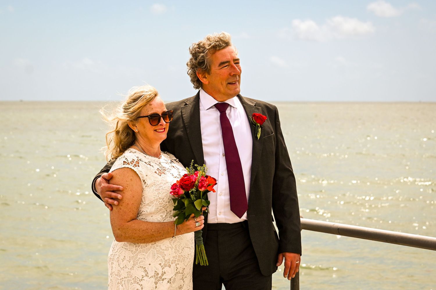 Couple in wedding attire smiling on a seaside pier, bride holding red roses, groom in dark suit with burgundy tie.