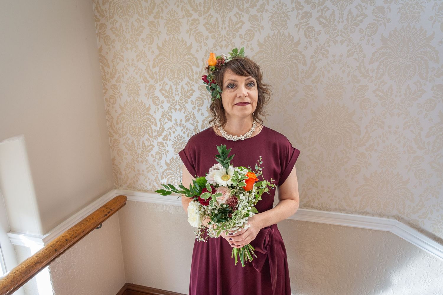 Woman in burgundy dress holding a floral bouquet with flowers in her hair, standing on a staircase.