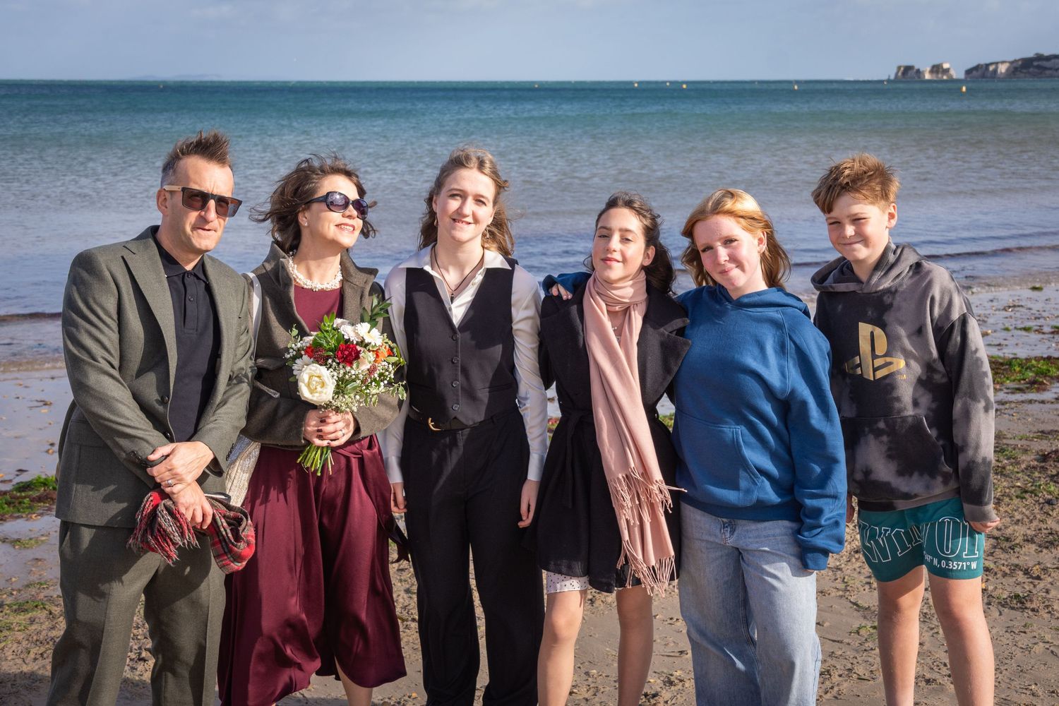 Family group photo on a beach, smiling together, one woman holding a bouquet in a burgundy dress.