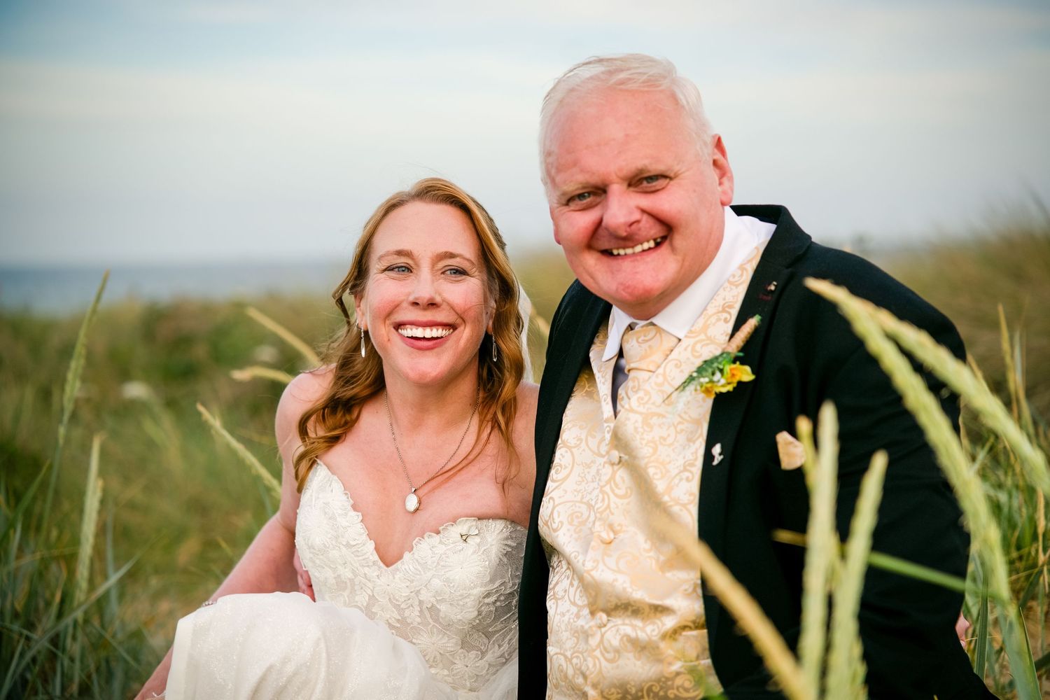 Happy couple posing outdoors in coastal grass, bride in lace gown and groom in tartan jacket on their wedding day.