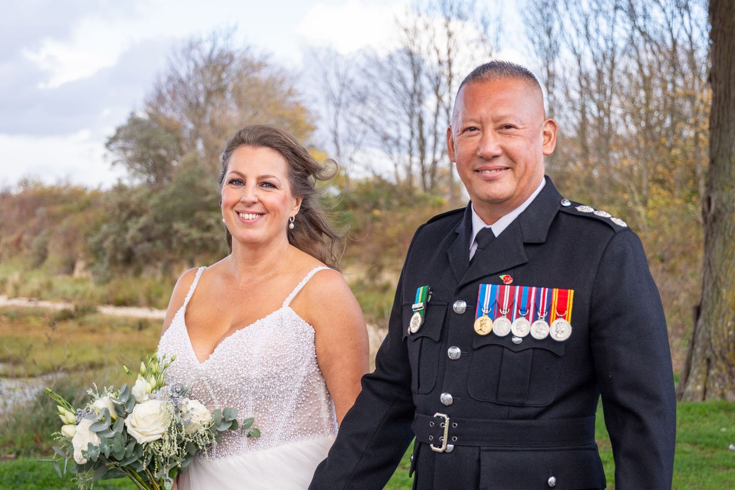 Bride in white gown holding bouquet stands with groom in military uniform displaying medals outdoors.