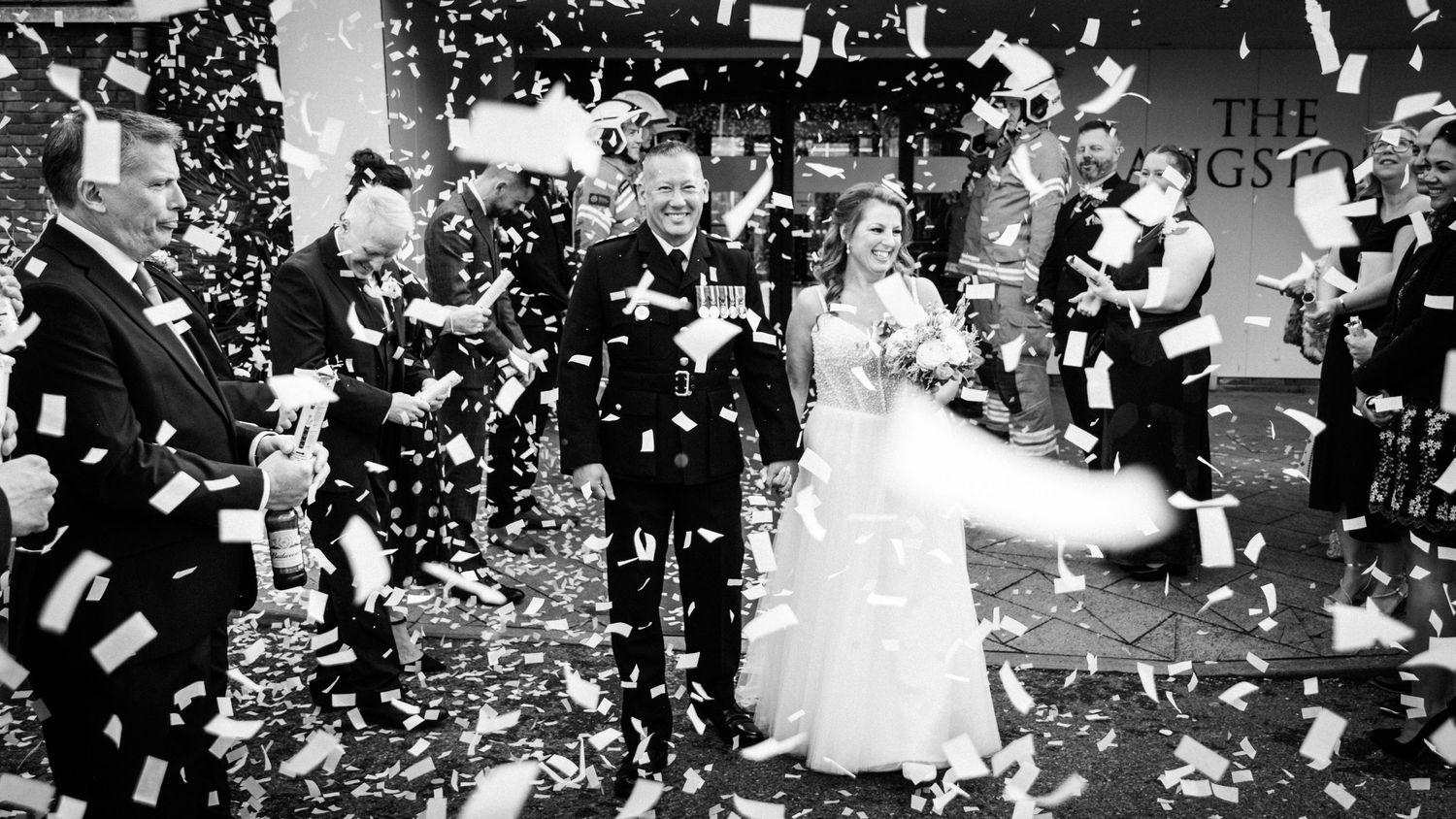 Black and white wedding photo of couple exiting ceremony through confetti shower with guests celebrating.