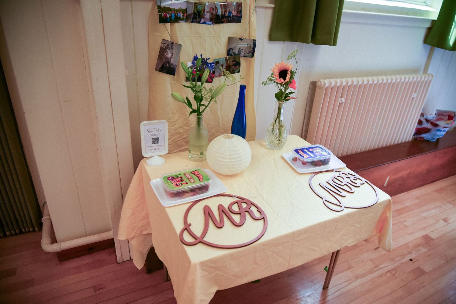 Wedding table with Mr and Mrs signs, flowers in vases, and photos displayed on a light pink tablecloth.
