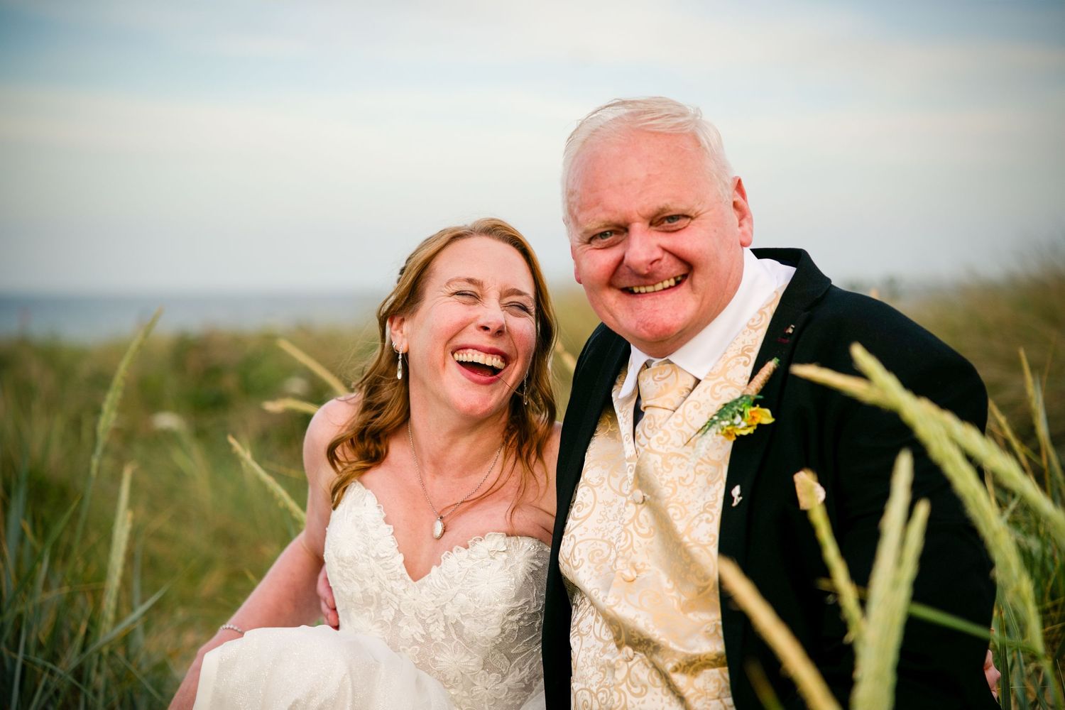 Joyful bride and groom laughing together outdoors near coastal grass dunes on their wedding day.