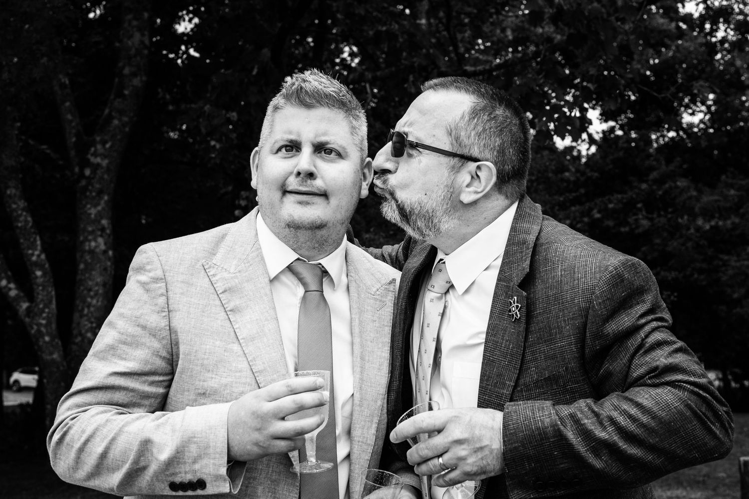 Two grooms share a kiss at their wedding reception outdoors in a black and white photo.