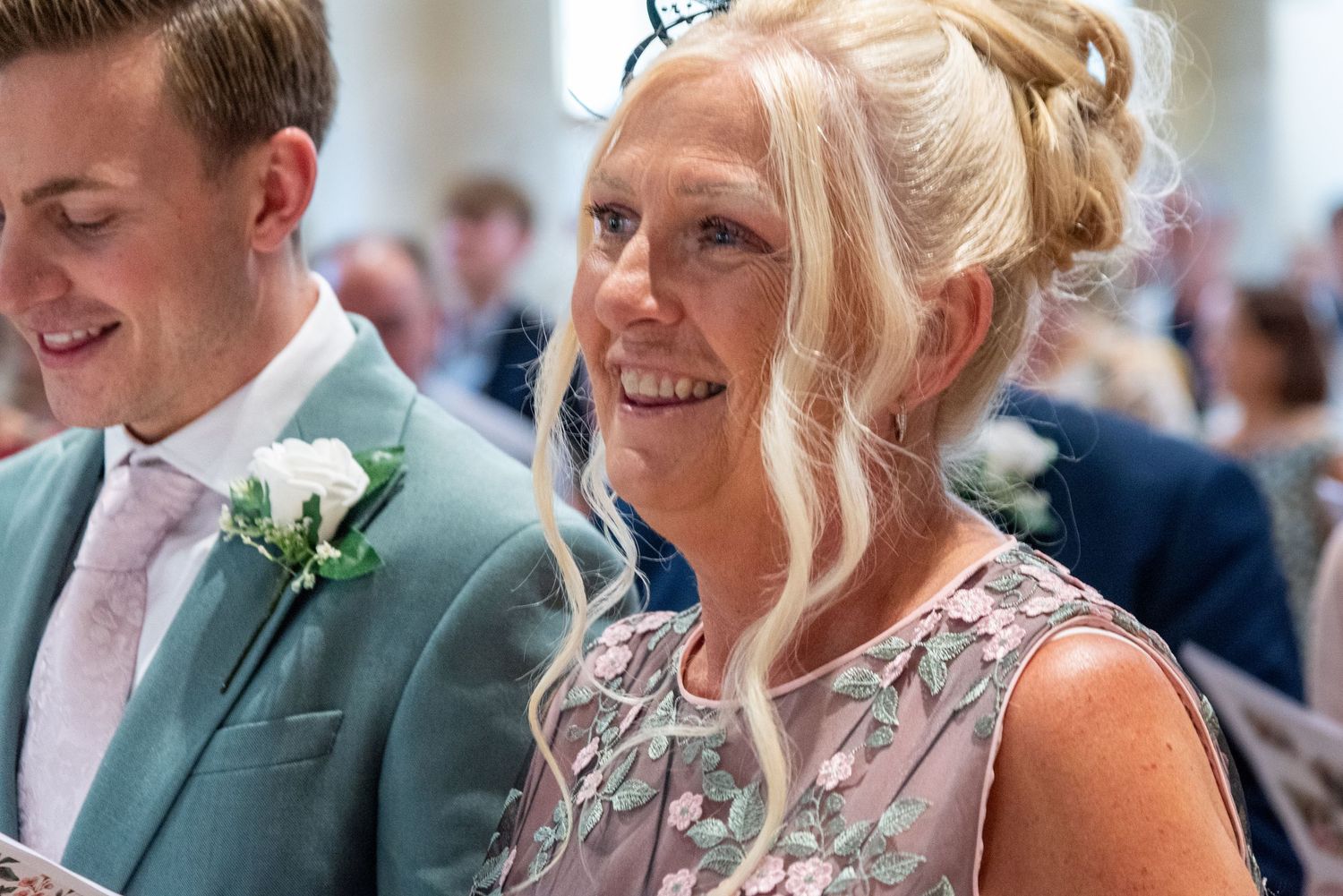 Smiling blonde woman in floral dress at wedding ceremony seated beside groom in mint suit.