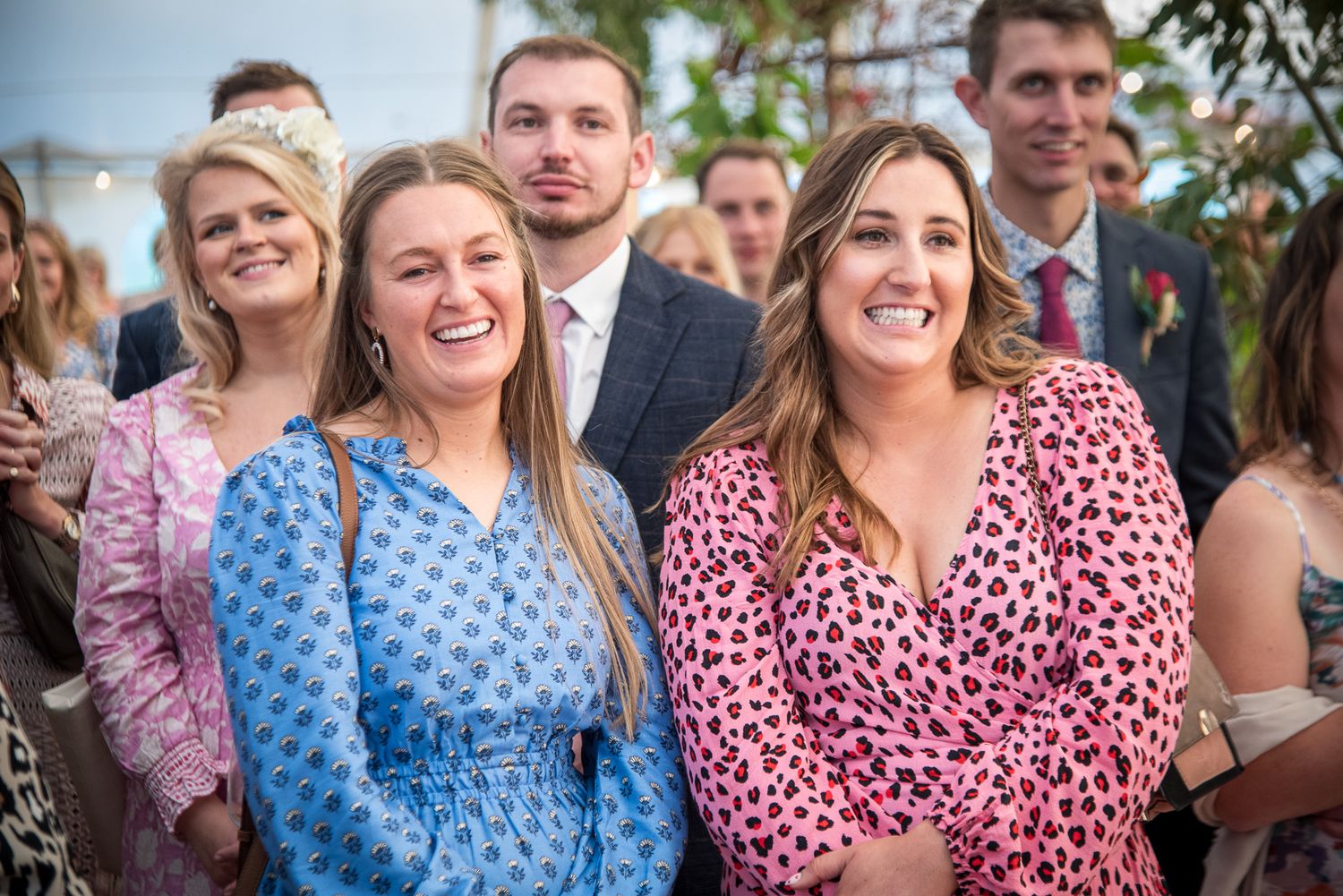 Two smiling women in floral dresses pose together at an outdoor wedding celebration with guests in background.