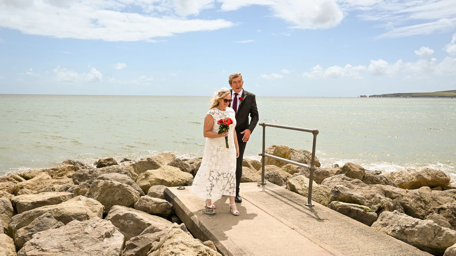 Bride and groom share a romantic moment on a rocky seaside pier, with turquoise ocean waters and blue skies behind them.