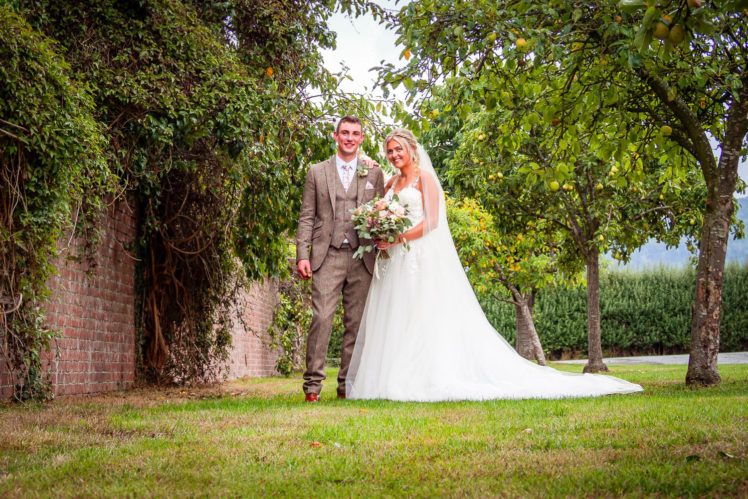 Bride and groom pose together in a lush garden setting, bride in white gown holding bouquet, groom in tan suit.