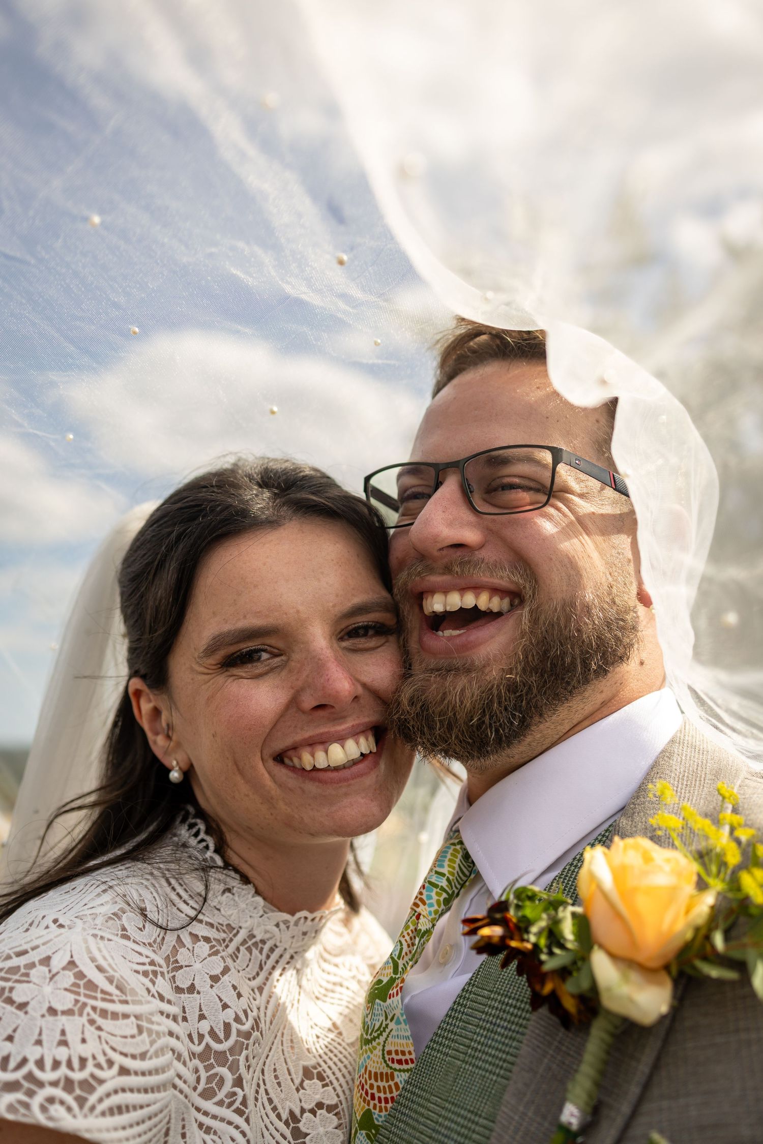 Happy bride and groom laughing together under a flowing wedding veil with blue sky backdrop.