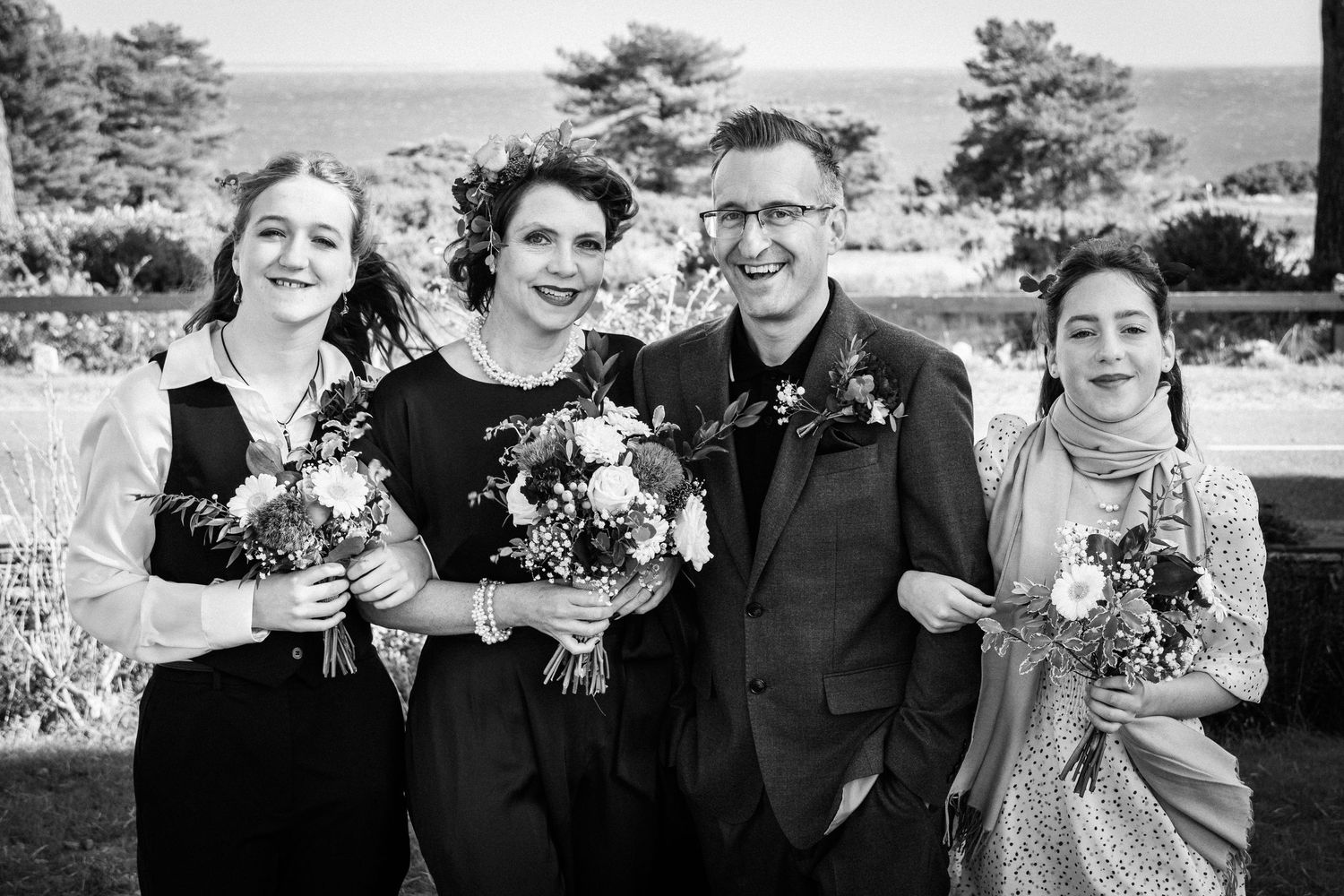 Black and white wedding photo of bride, groom, and two bridesmaids holding bouquets outdoors near the ocean.