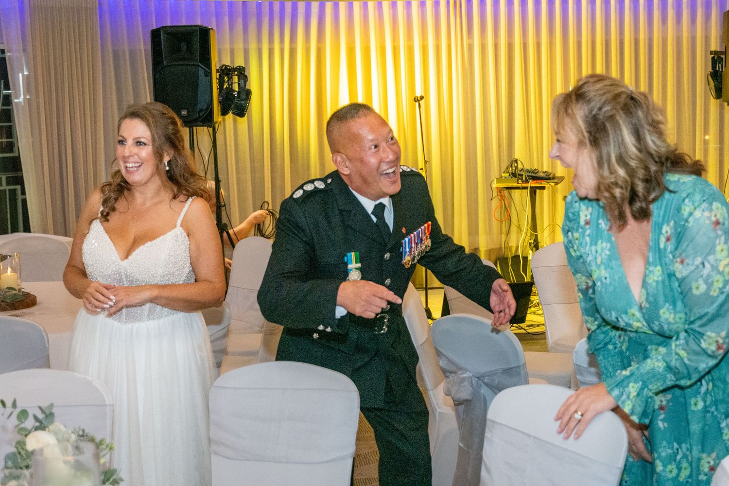 Military officer in uniform laughing with two women in elegant attire at a formal wedding reception.