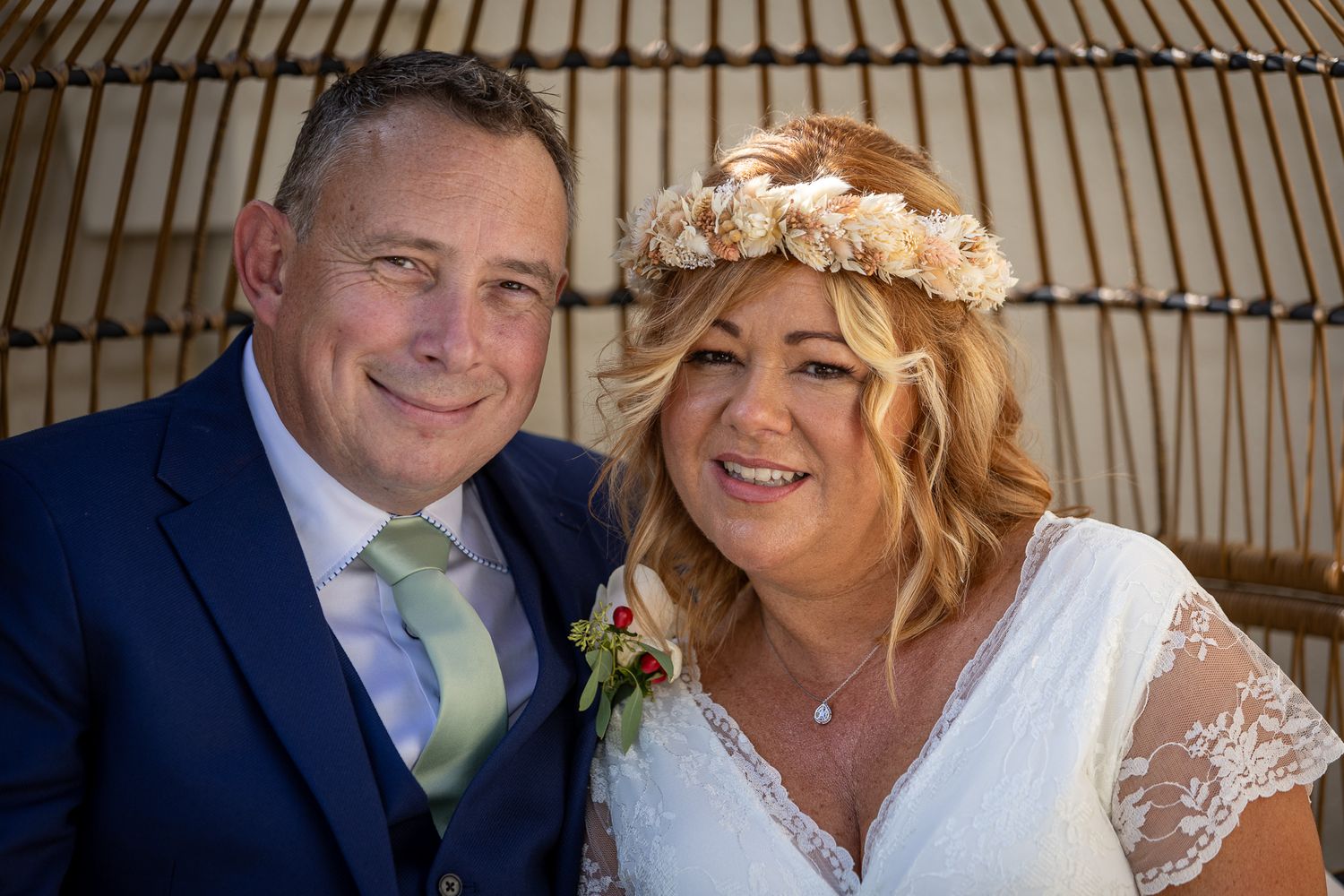 A smiling couple in wedding attire pose together, the bride wearing a floral crown and lace dress, the groom in a navy suit with green tie.