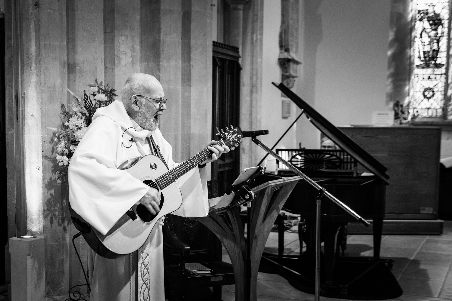 Elderly priest in white vestments playing acoustic guitar at church altar in black and white photo.