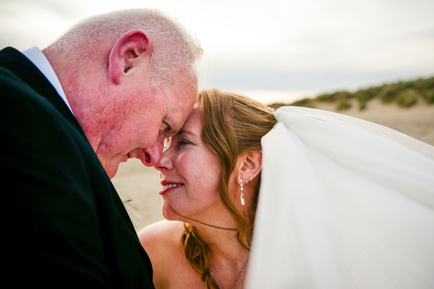 Bride and groom share an intimate forehead touch on a beach, bride wearing veil, in a romantic wedding portrait.