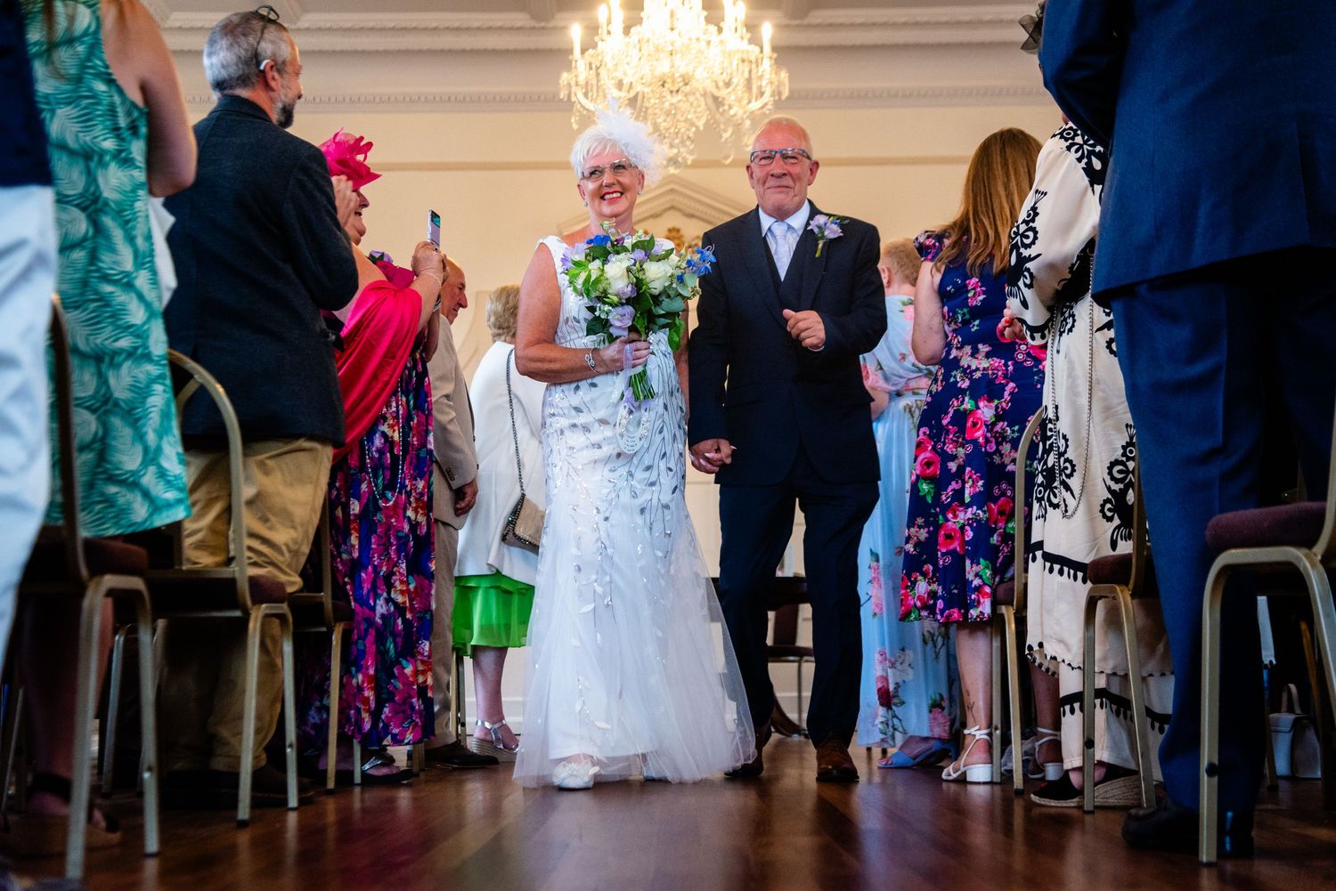 Elderly bride and groom walk down the aisle at elegant wedding ceremony, guests applauding in decorated ballroom.
