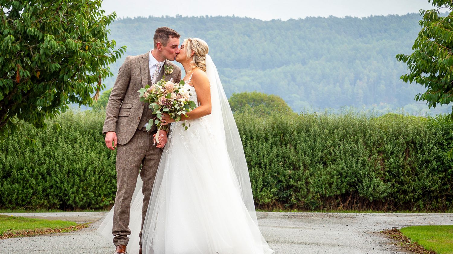 Bride and groom share a romantic kiss outdoors on a rainy wedding day with lush green hills in the background.