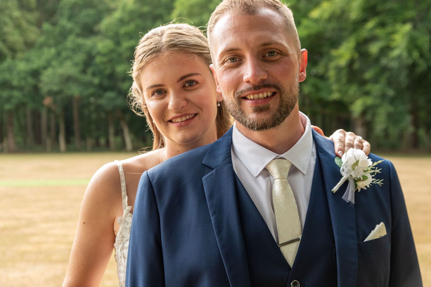 A couple poses outdoors at their wedding, with the groom wearing a navy blue suit and the bride in white against a wooded background.