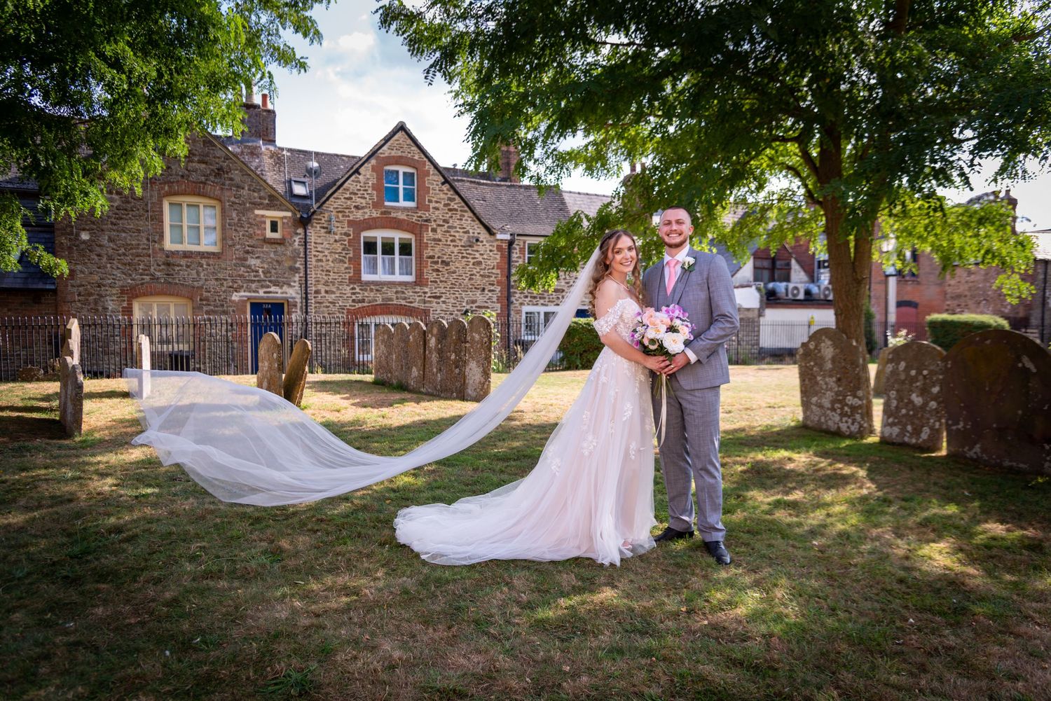 Bride and groom pose outside a stone church with veil flowing in the breeze.