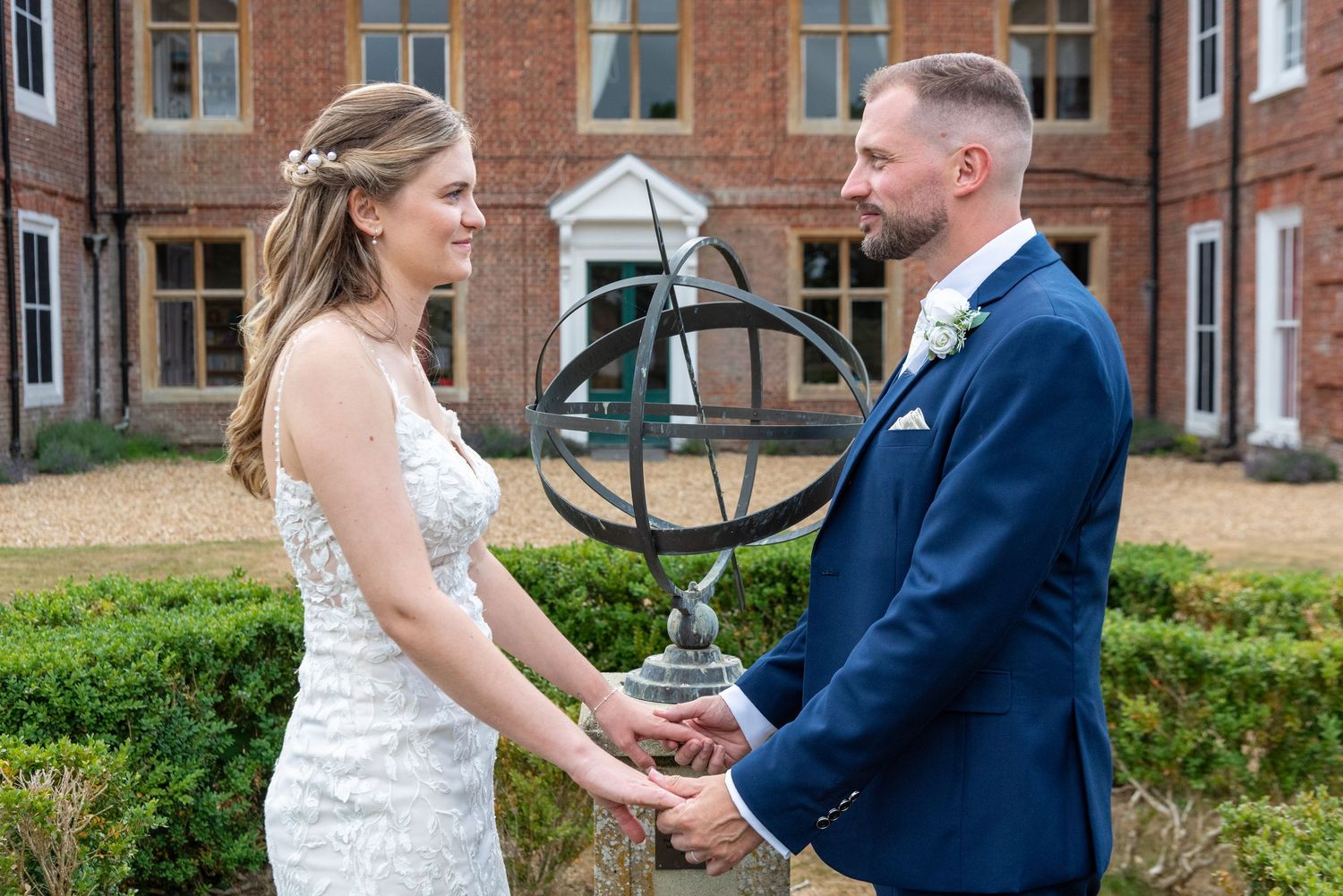 Bride and groom holding hands in a formal garden with sundial, groom in navy suit, bride in lace gown.