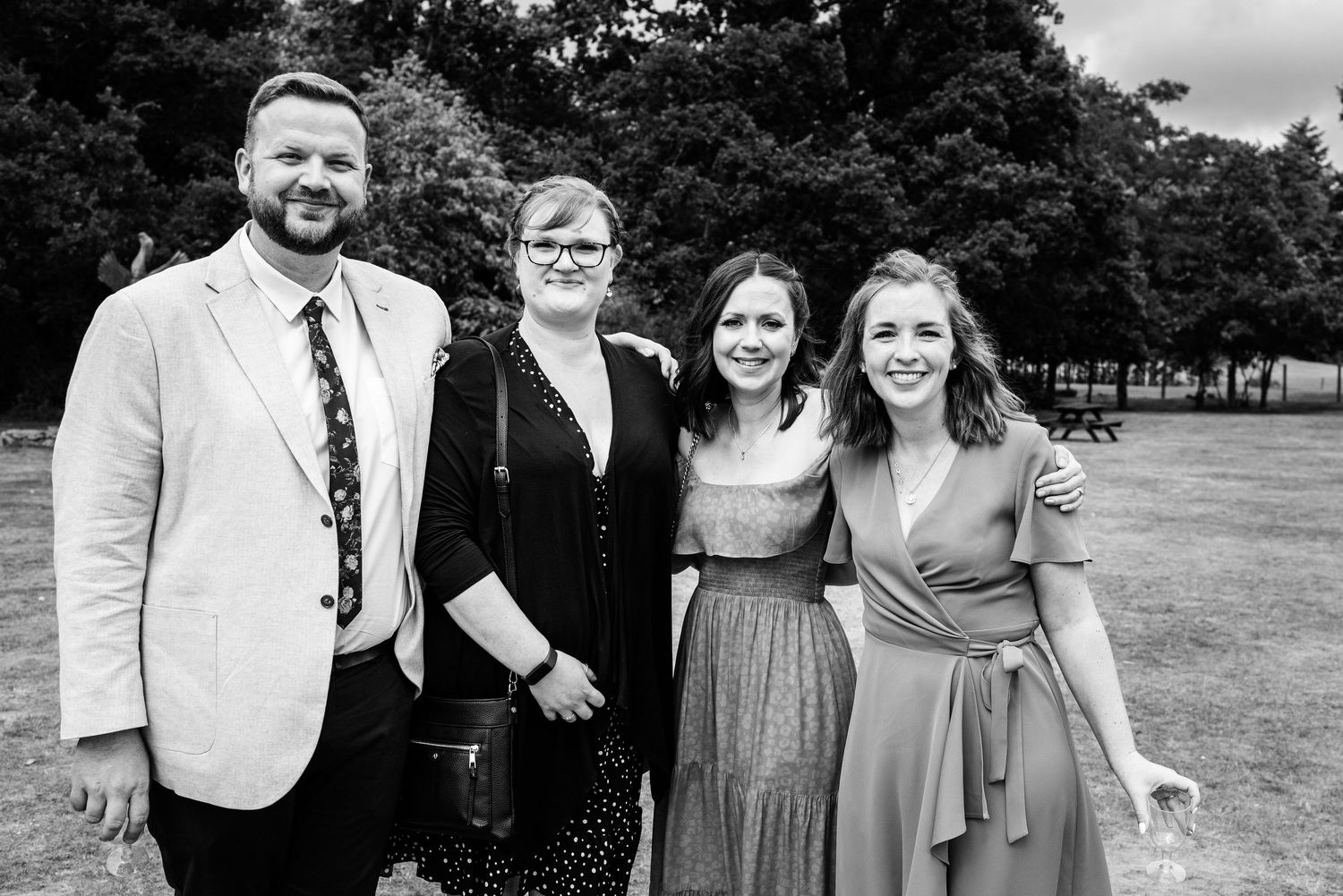 Black and white photo of four wedding guests smiling together outdoors on a sunny day.