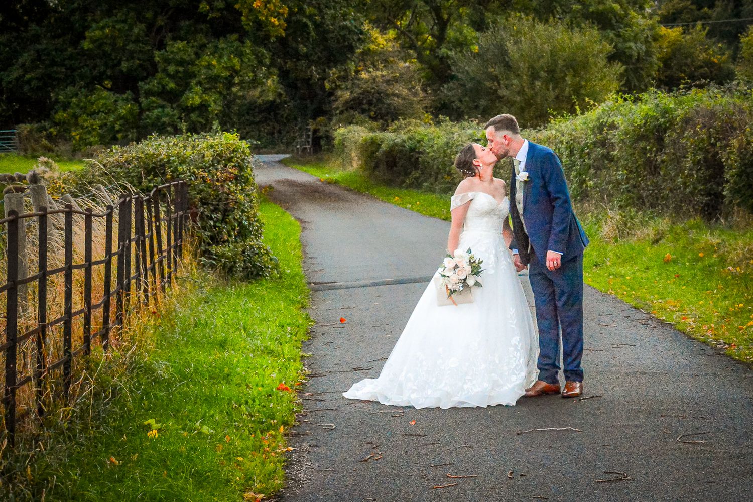 Bride and groom sharing a kiss on a scenic path surrounded by lush greenery on their wedding day.
