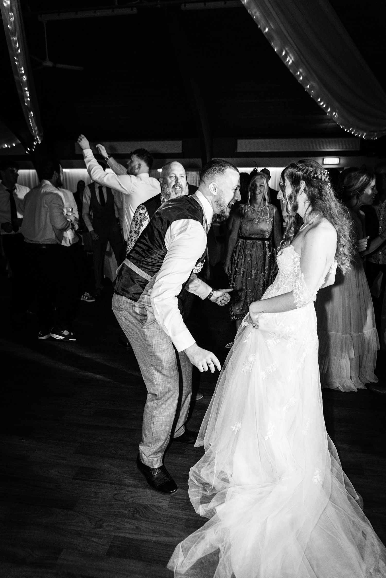 Black and white photo of bride and groom dancing joyfully at their wedding reception with guests in background.