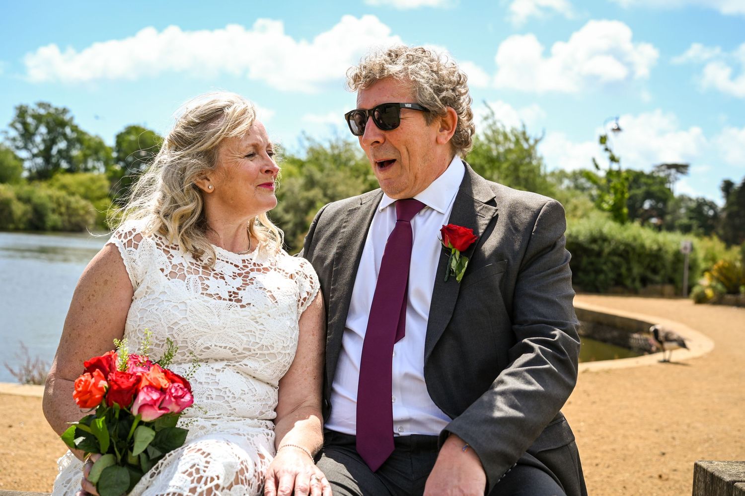 Bride and groom laughing together outdoors near a lake on their wedding day, bride holding colorful bouquet.