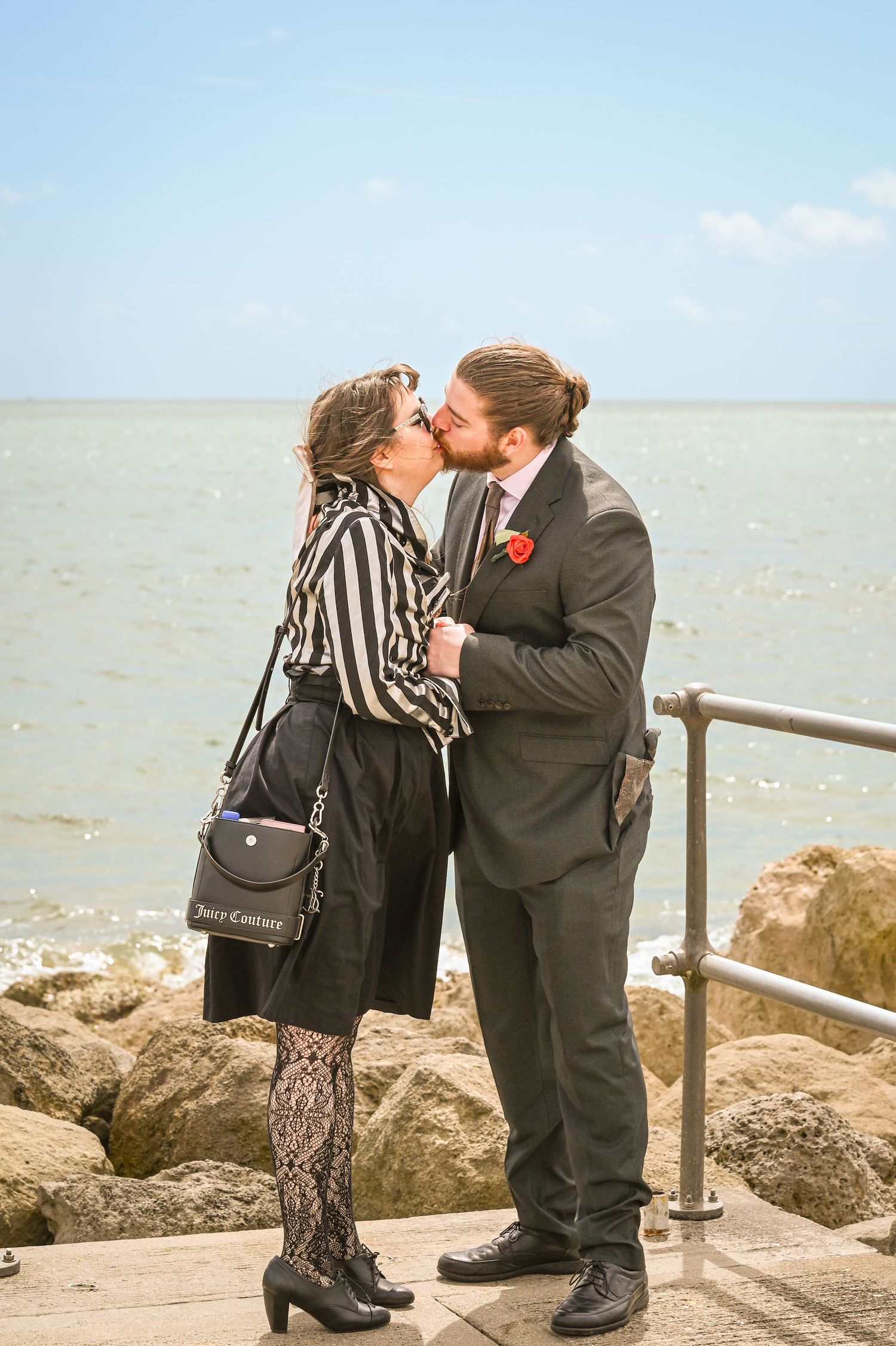 Couple kissing romantically by the seaside, woman in striped blouse with Juicy Couture bag, man in dark suit with red boutonniere.