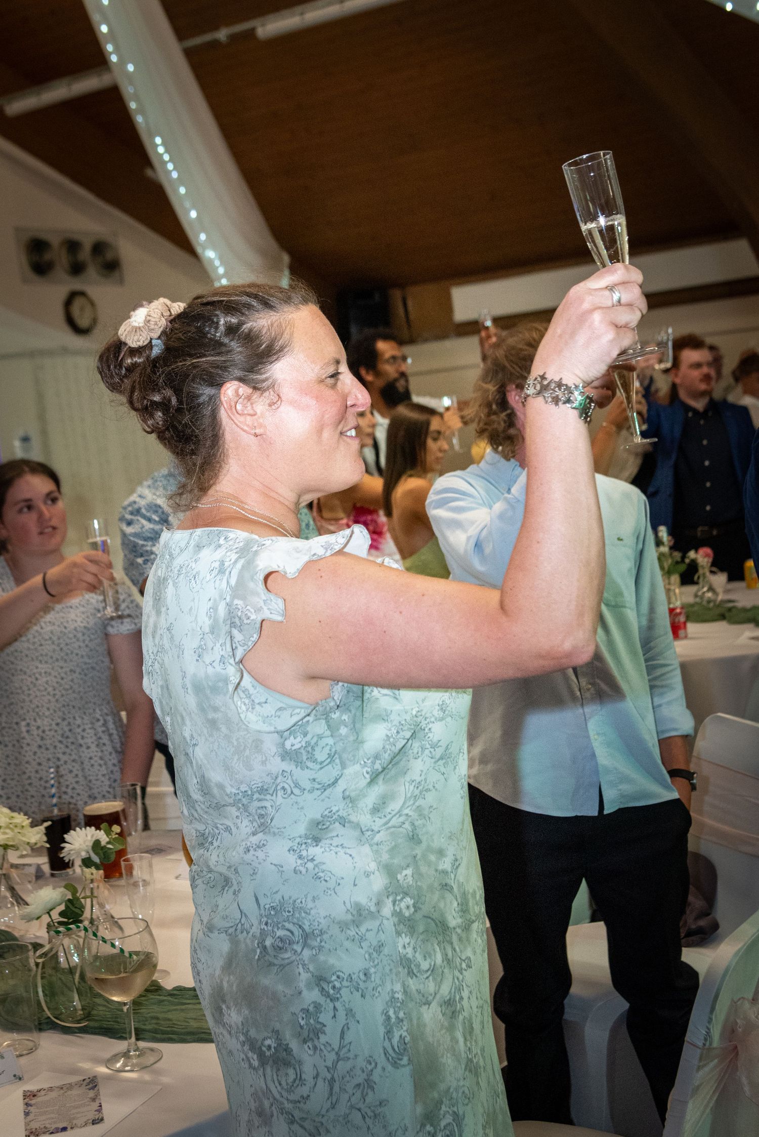 Woman in sage green dress raising a champagne flute during a celebratory wedding reception toast.