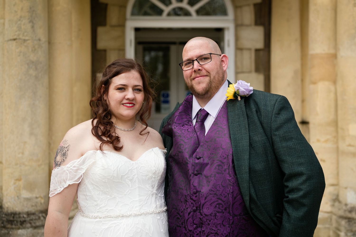 Bride and groom posing together outside a stone building on their wedding day, wearing white dress and green suit.