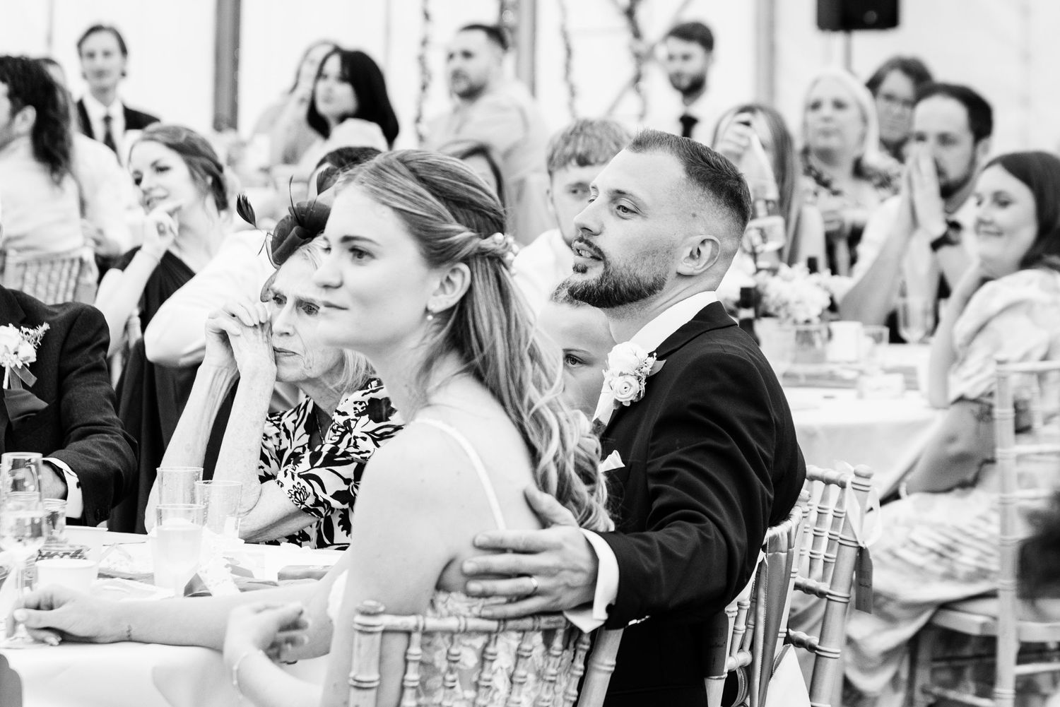 Black and white wedding photo of bride and groom sitting together at reception table surrounded by guests.