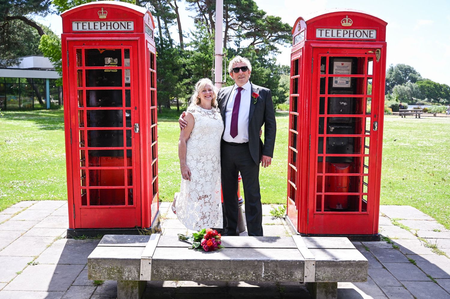 Couple posing between two classic red British telephone boxes outdoors on a sunny day at what appears to be a wedding.