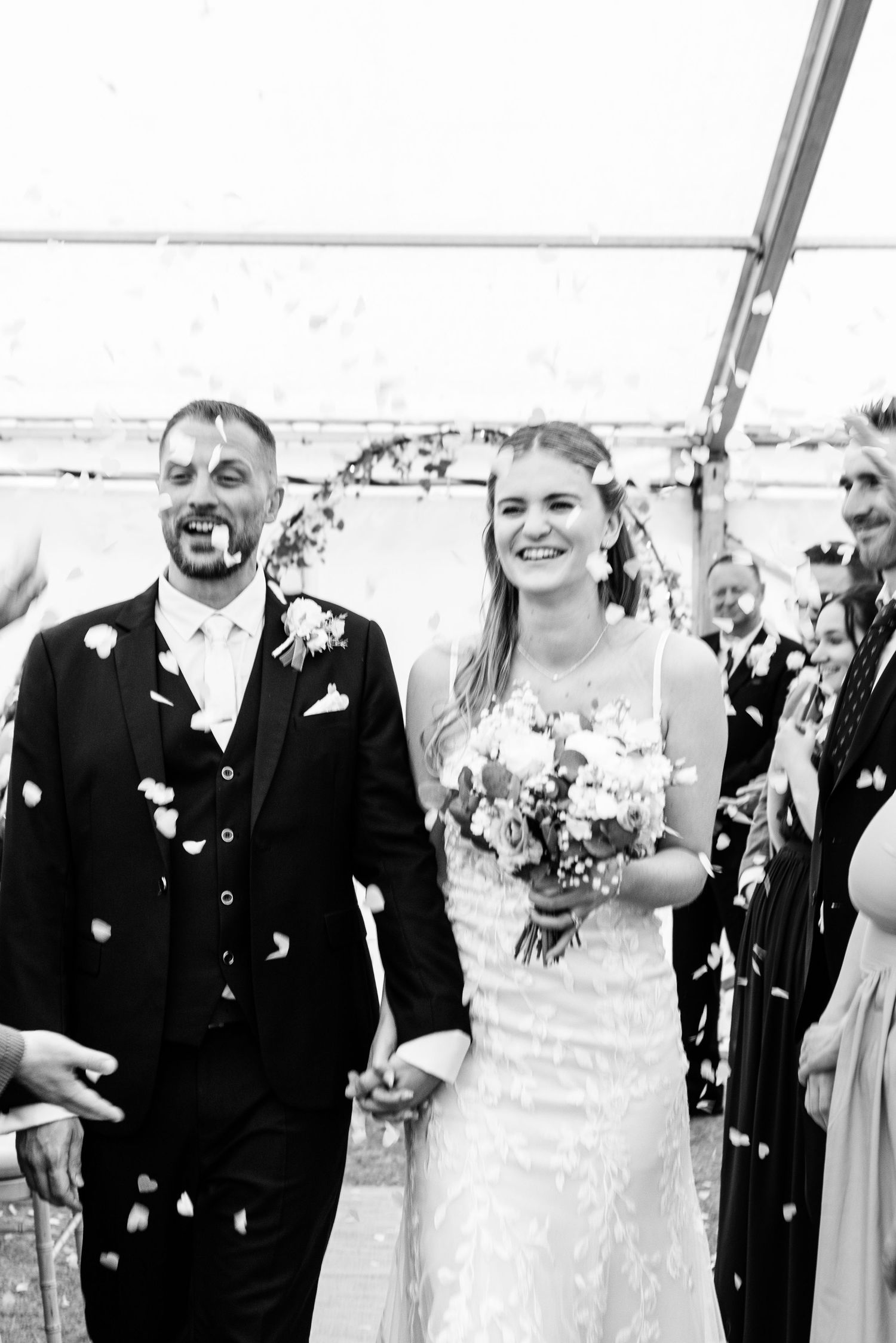 Black and white photo of smiling bride and groom walking through confetti shower.