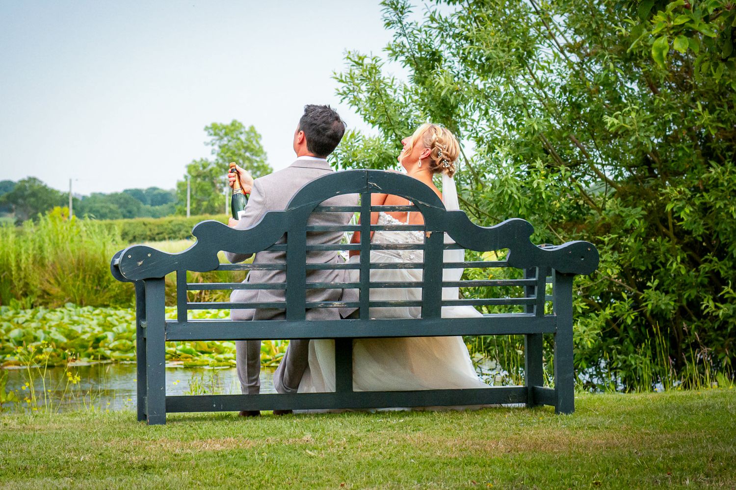 Couple in wedding attire sitting together on a green garden bench overlooking a serene pond surrounded by lush greenery.