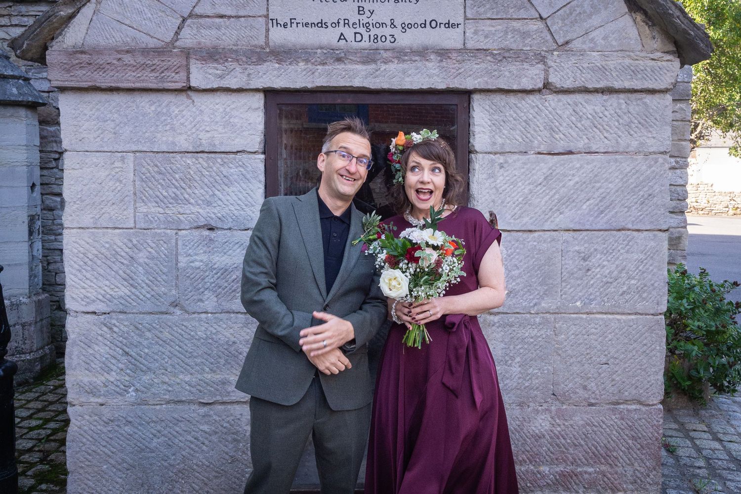 Couple smiling in front of stone monument, woman in burgundy dress holding floral bouquet, man in gray suit.