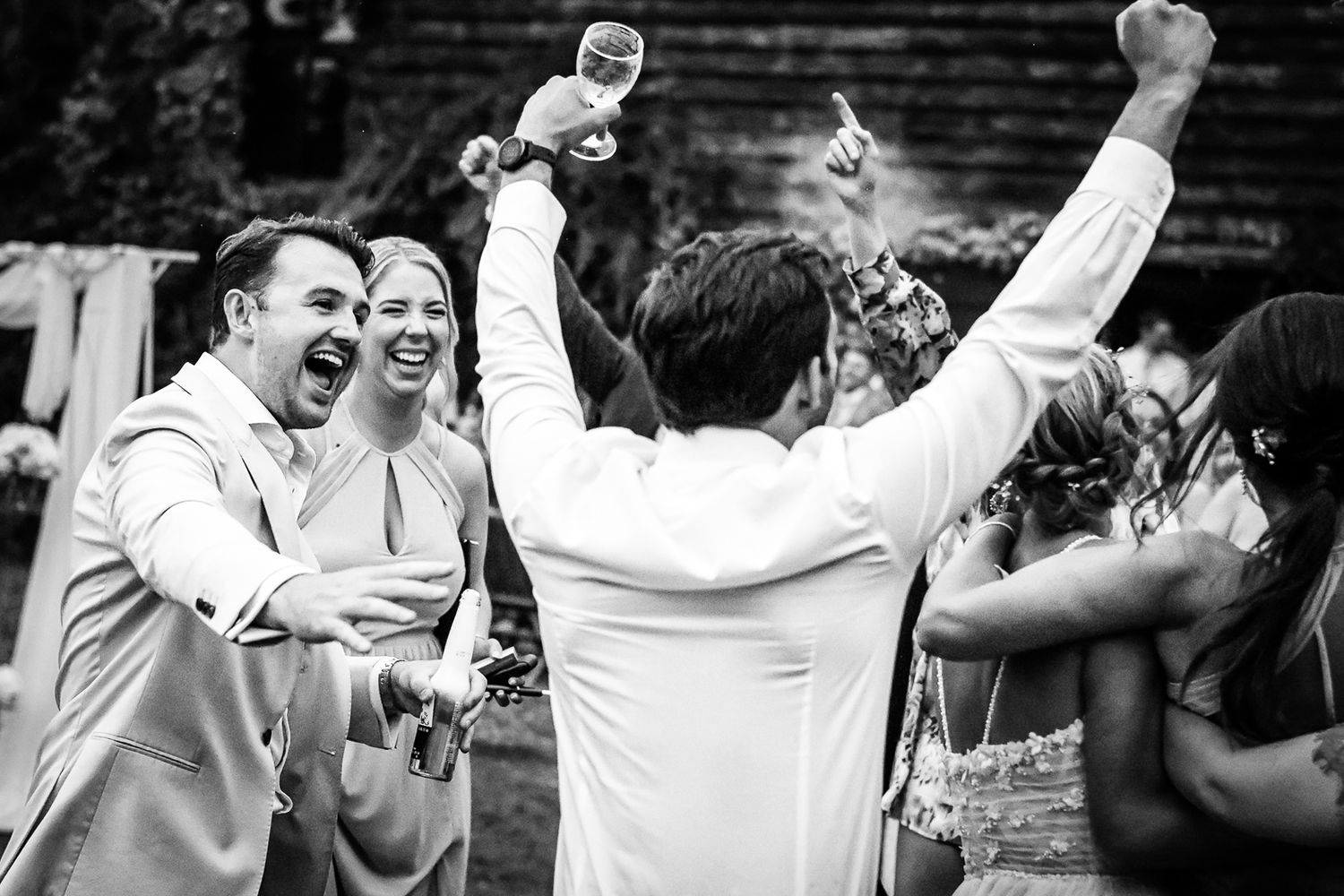Black and white wedding celebration photo of joyful guests cheering and raising glasses outdoors.