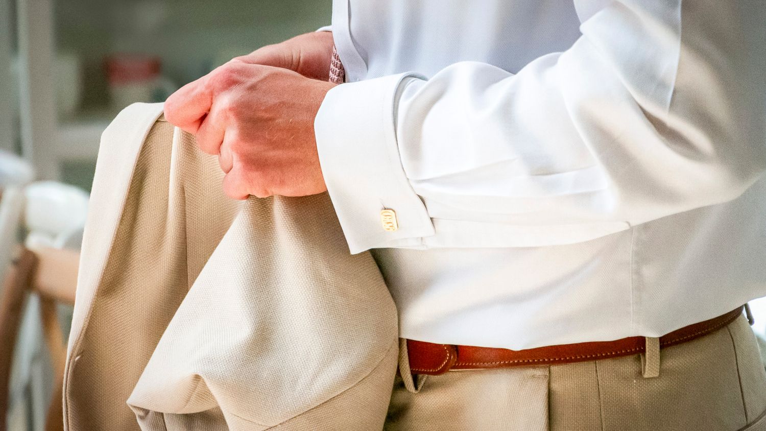 Man in white dress shirt holding a beige suit jacket, wearing brown leather belt and tan trousers.