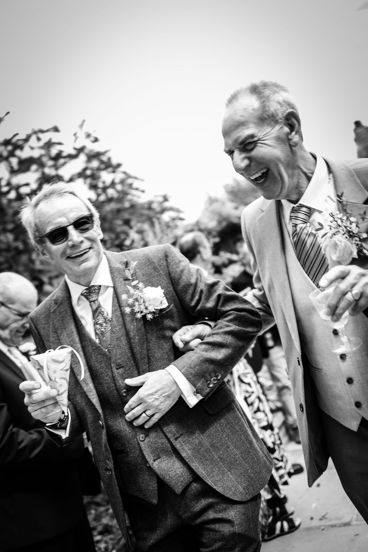 Two grooms laugh joyfully together during wedding confetti celebration in black and white photo.