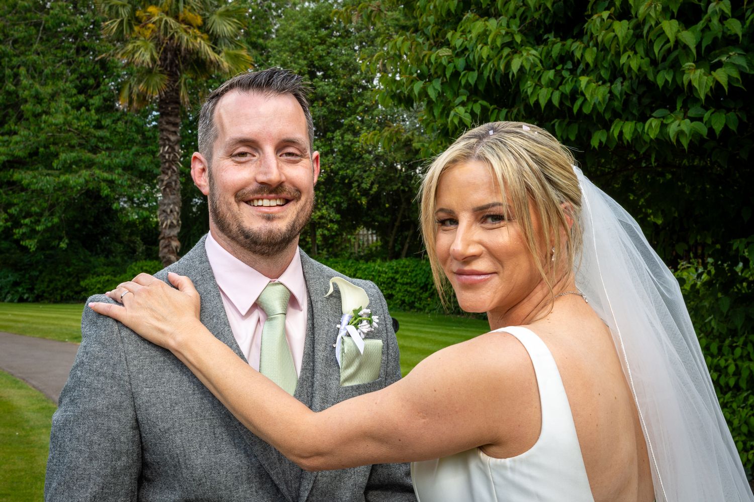 Happy couple posing outdoors on their wedding day, bride in white gown with veil, groom in grey suit with pink tie.