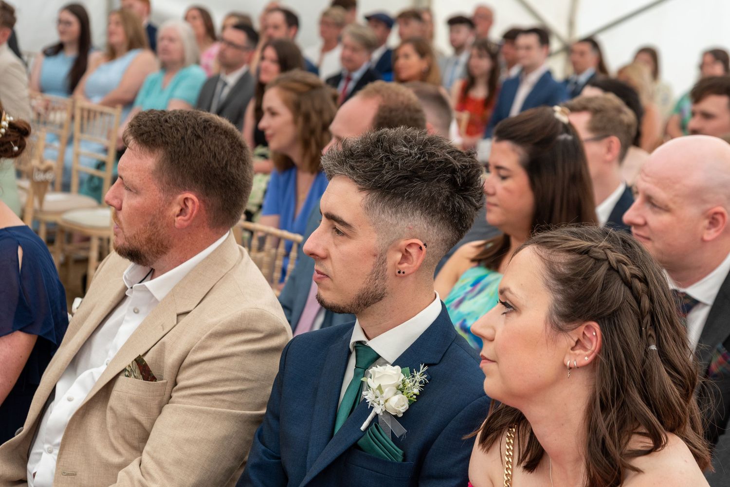 Wedding guests seated in rows attentively watching a ceremony inside a bright marquee tent.