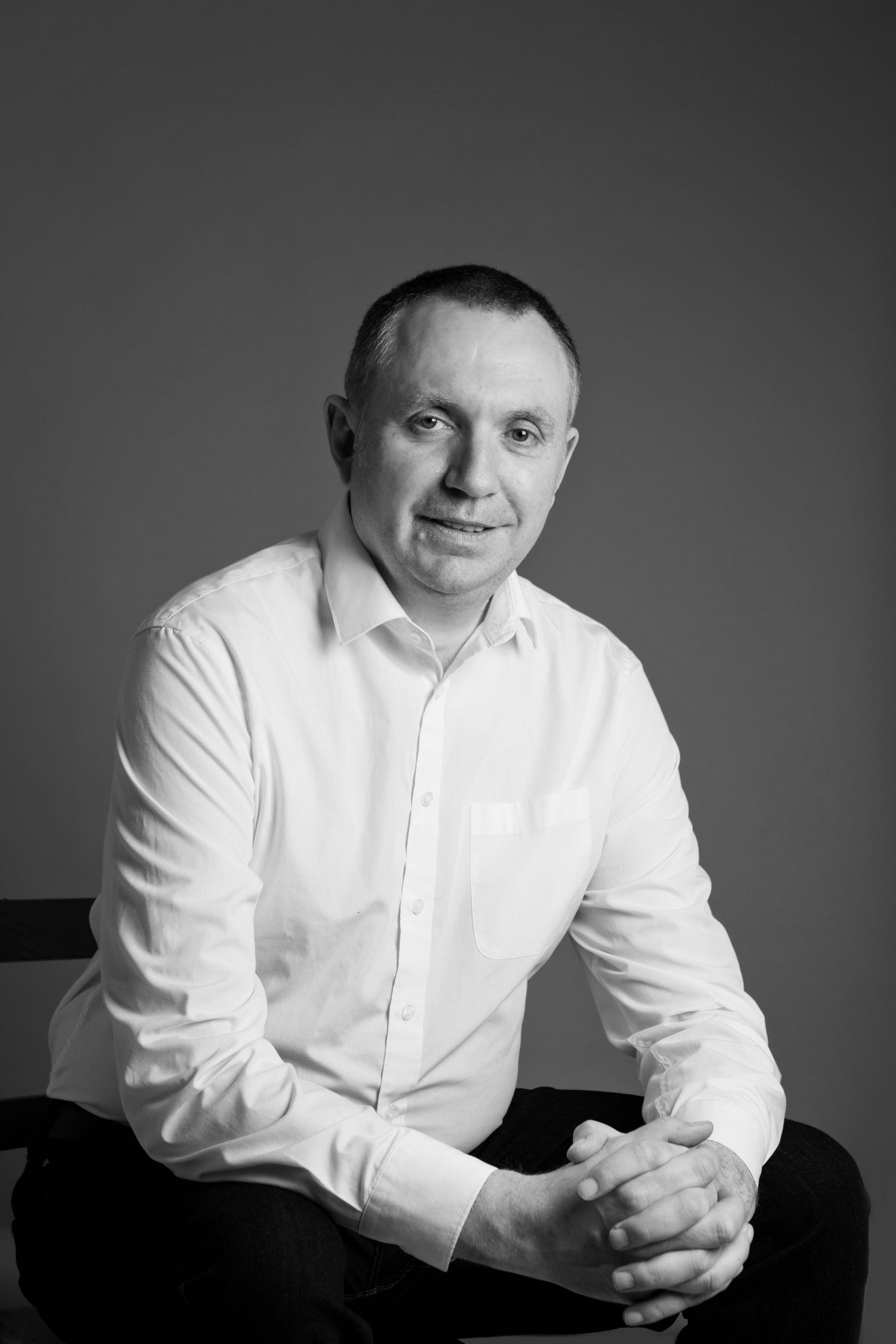 Black and white studio portrait of smiling man in white shirt seated with hands clasped.